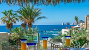 A balcony with a table set for two, overlooking a beach with palm trees and a clear blue sea.