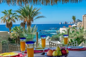 A balcony with a table set for two, overlooking a beach with palm trees and a clear blue sea.