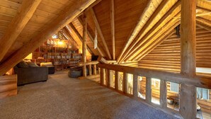 A spacious attic room with a sloped wooden ceiling, a cozy sofa, a bookshelf, and a view through the windows. - Rowland Log Cabin 3 Bedroom Home by RedAwning (Lyle)