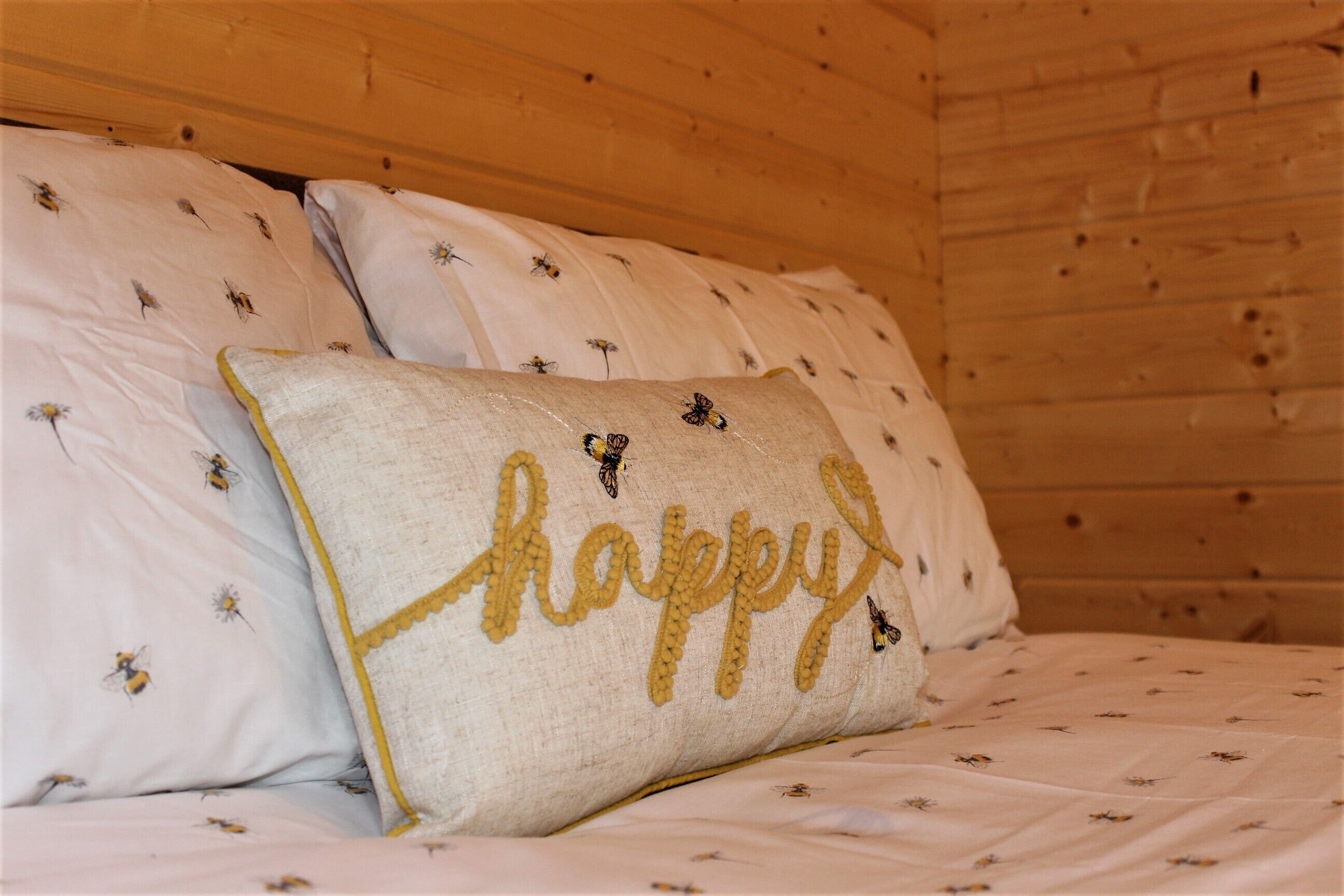 A bed with white bedding featuring small bee patterns and a pillow with the word "happy" embroidered in yellow.