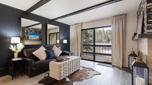 A modern living room with a dark wooden ceiling, a large window with a view of trees, a leather sofa with cushions, a small table with a lamp, and a glass fireplace. - Luxurious 2 Bedroom Base Camp Condo - Steps from Kirkwood Village 2 Condo by RedAwning (Kirkwood)