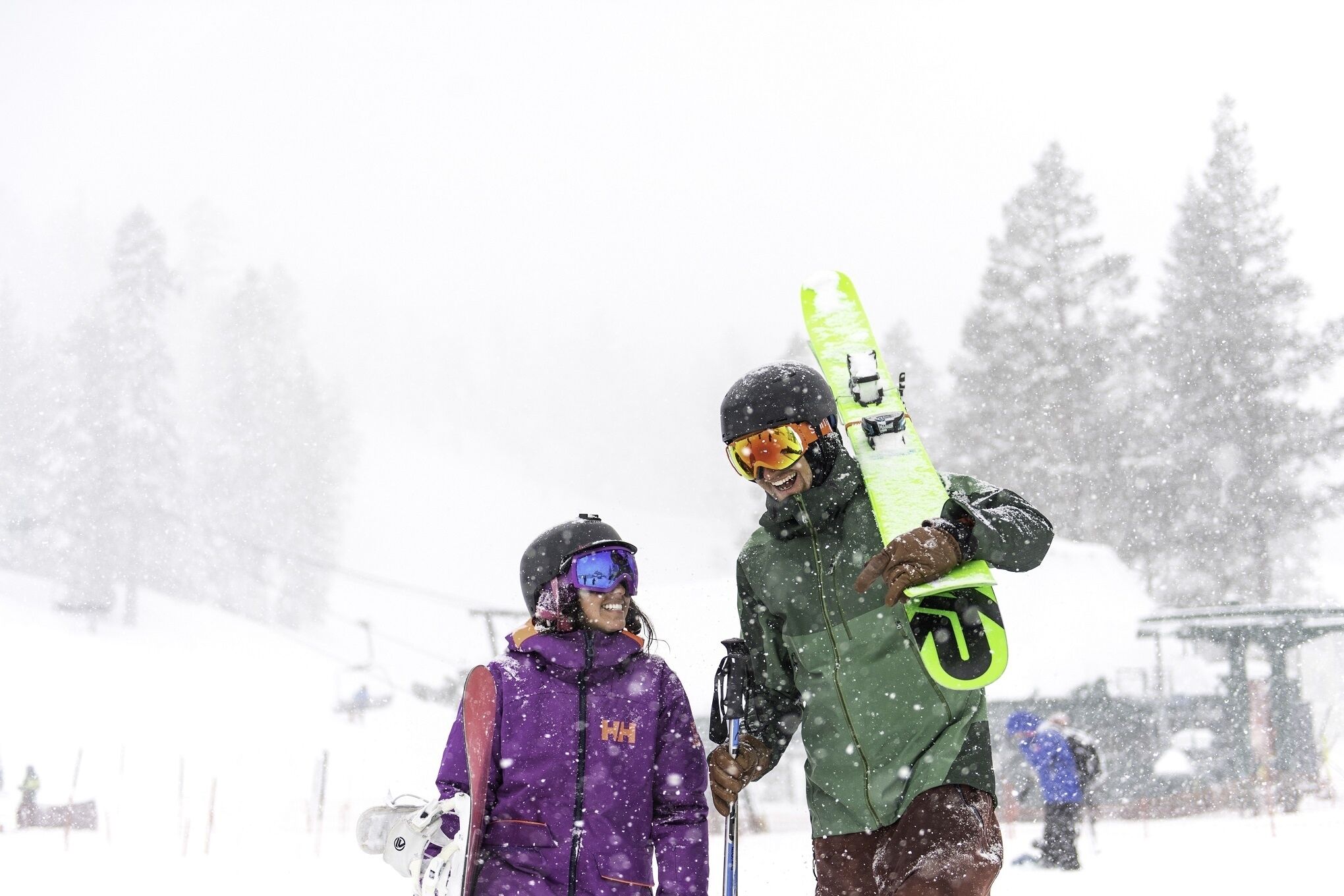 Two people with skis, one holding a neon green ski, standing in a snowy area with trees and a ski lift in the background.