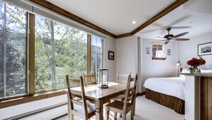 A room with a dining table, chairs, a bed, a ceiling fan, and large windows with a view of trees. - Spacious Studio Condo With Lift View At Elkhorn Lodge 1 Bedroom Condo by RedAwning (Avon)