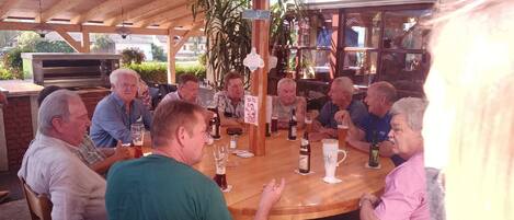 Un groupe de personnes est assis autour d’une table en bois, dans un cadre extérieur. On aperçoit un plafond en bois et un four à pizza à l’arrière-plan.