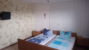 Quarto com cama de madeira, mesa de cabeceira, televisão fixada na parede e uma cruz na parede.