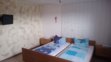 Quarto com cama de madeira, mesa de cabeceira, televisão fixada na parede e uma cruz na parede.