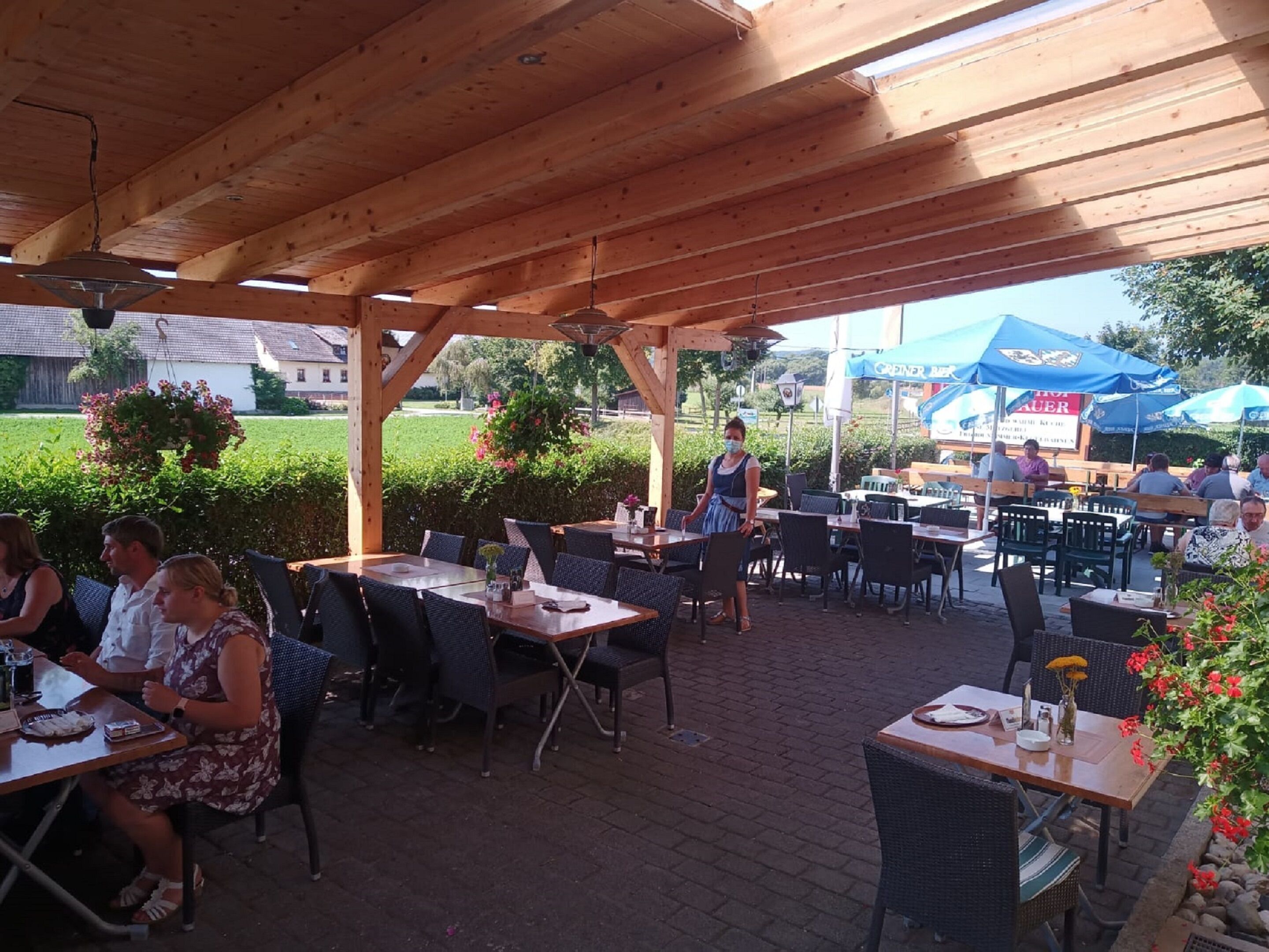 An outdoor patio with wooden tables and chairs, surrounded by greenery and a clear sky.