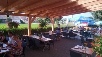 An outdoor patio with wooden tables and chairs, surrounded by greenery and a clear sky.