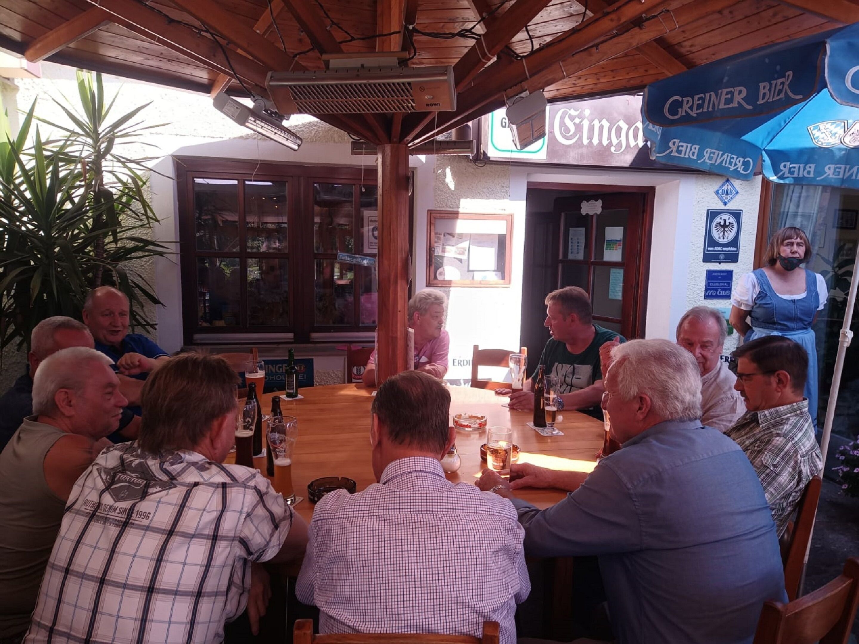 Een groep mensen zit rond een houten tafel in een restaurant met een houten plafond en een bord waarop 'Greiner Bier' staat.