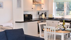 A modern kitchen with white cabinetry, a black microwave, and a dining area with white chairs and a table set for two. - Suncroft (Port Isaac)
