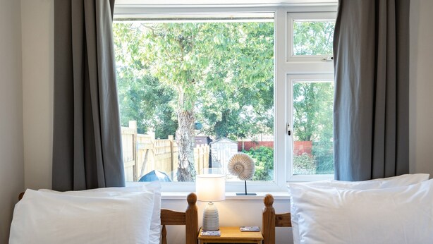 A bedroom with two beds, a window with curtains, a bedside table with a lamp, and a view of trees and a fence outside. - Suncroft (Port Isaac)