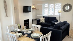 A dining area with a round table set for four, surrounded by white chairs. A flat-screen TV is mounted on the wall above the table. - Suncroft (Port Isaac)
