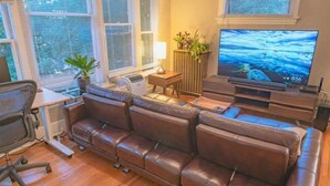 A living room with a brown leather sofa, a flat-screen TV, a small table with a lamp, and a potted plant.