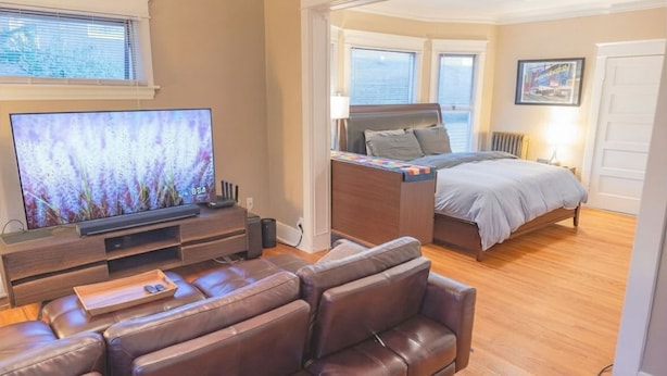 A modern living room with a flat-screen TV, leather sofa, and a bed with a gray headboard.