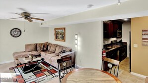 A living room with a beige sofa, a coffee table, and a dining table with chairs. There is a ceiling fan, a clock, and a painting on the wall. - Beach Block Retreat w/Patio, Off Street Parking, Linens & Towels, Beach Gear (Seaside Heights)
