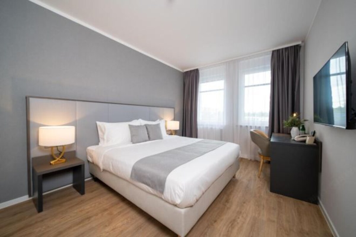 A modern hotel room with a large bed, bedside tables with lamps, a desk, and a flat-screen TV mounted on the wall.