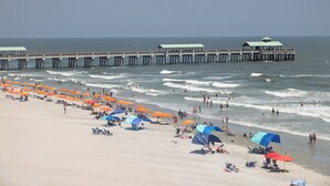 Une plage avec une jetée qui s’avance dans l’océan, des parasols et des gens qui se détendent sur le sable.