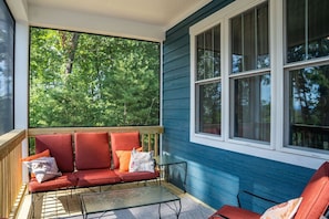 A covered porch with a red sofa and armchairs, a small table, and a window with a view of trees.