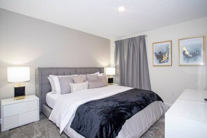 Une chambre moderne avec un grand lit, des tables de chevet avec des lampes, une commode blanche et deux tableaux abstraits encadrés accrochés au mur.