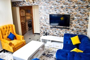 A living room with a TV mounted on a brick-patterned wall, a yellow armchair, a blue sofa, and a white coffee table.