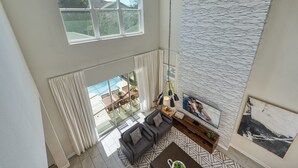 Ein Wohnzimmer mit hohen Decken, einem großen Fenster mit Blick auf einen Pool, einem grauen Sofa und einem an der Wand befestigten Fernseher.