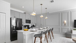 A modern kitchen with white cabinetry, a central island with bar stools, stainless steel appliances, pendant lighting, and a flat-screen TV mounted on the wall.