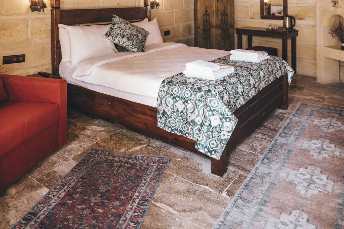Un dormitorio con techo de madera, cama con ropa de cama estampada, una silla roja, mesita de noche de madera y pared de piedra.