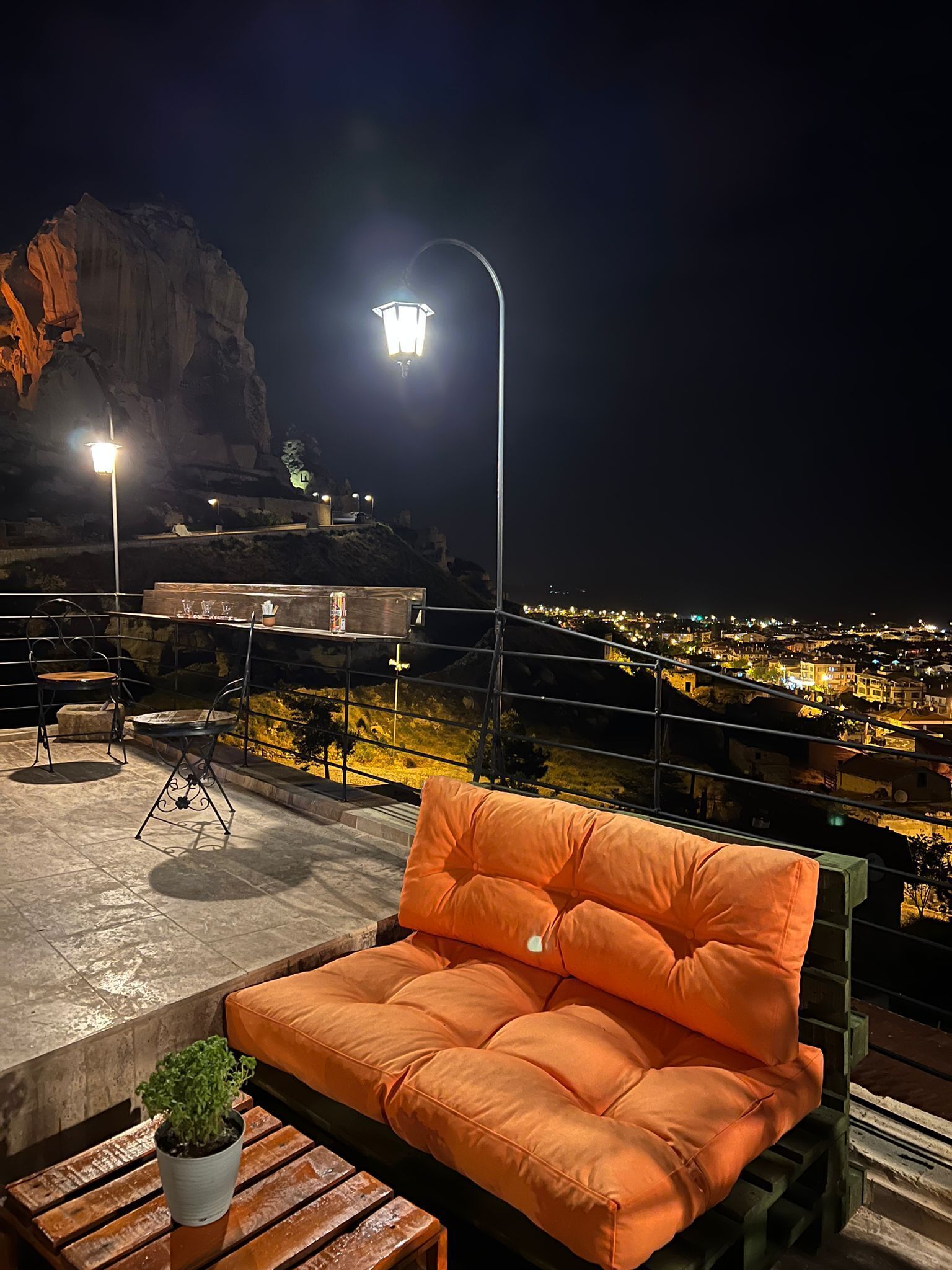Um terraço no telhado com área de estar acolchoada, uma pequena mesa com uma planta e vista para a cidade à noite.