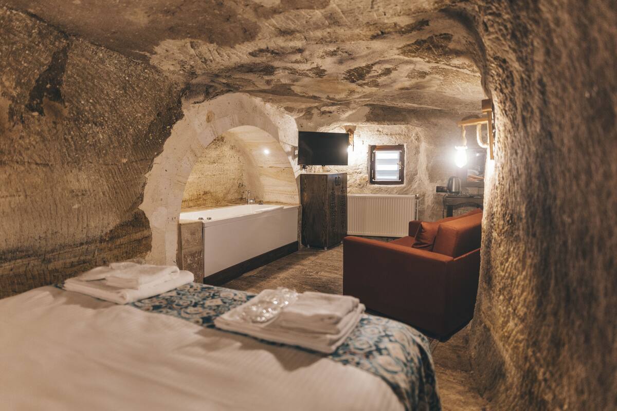 Una habitación acogedora con paredes de piedra, una cama, un sillón rojo, una mesita con un hervidor y un televisor montado en la pared.