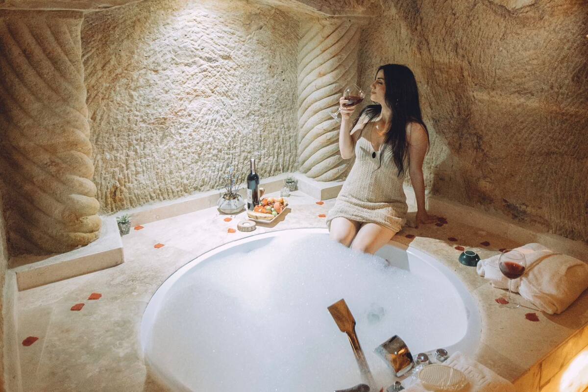 Una mujer sentada en una bañera en un ambiente que recuerda a una cueva, con una botella de vino y pétalos de rosa.