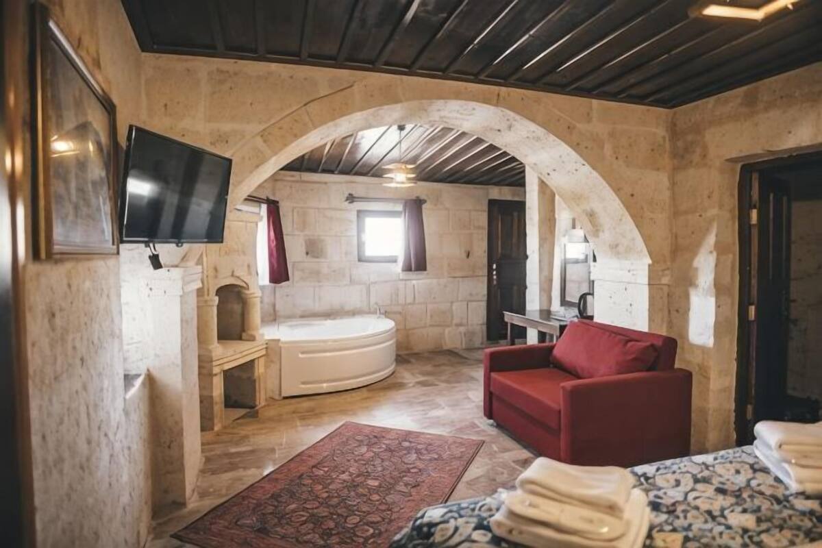 Una habitación de paredes de piedra con un sofá rojo, una bañera blanca y un televisor montado en la pared.