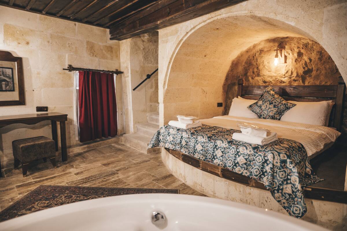 Un dormitorio de paredes de piedra con un amplio ventanal de arco, una cama de madera, una mesita de noche y una bañera.