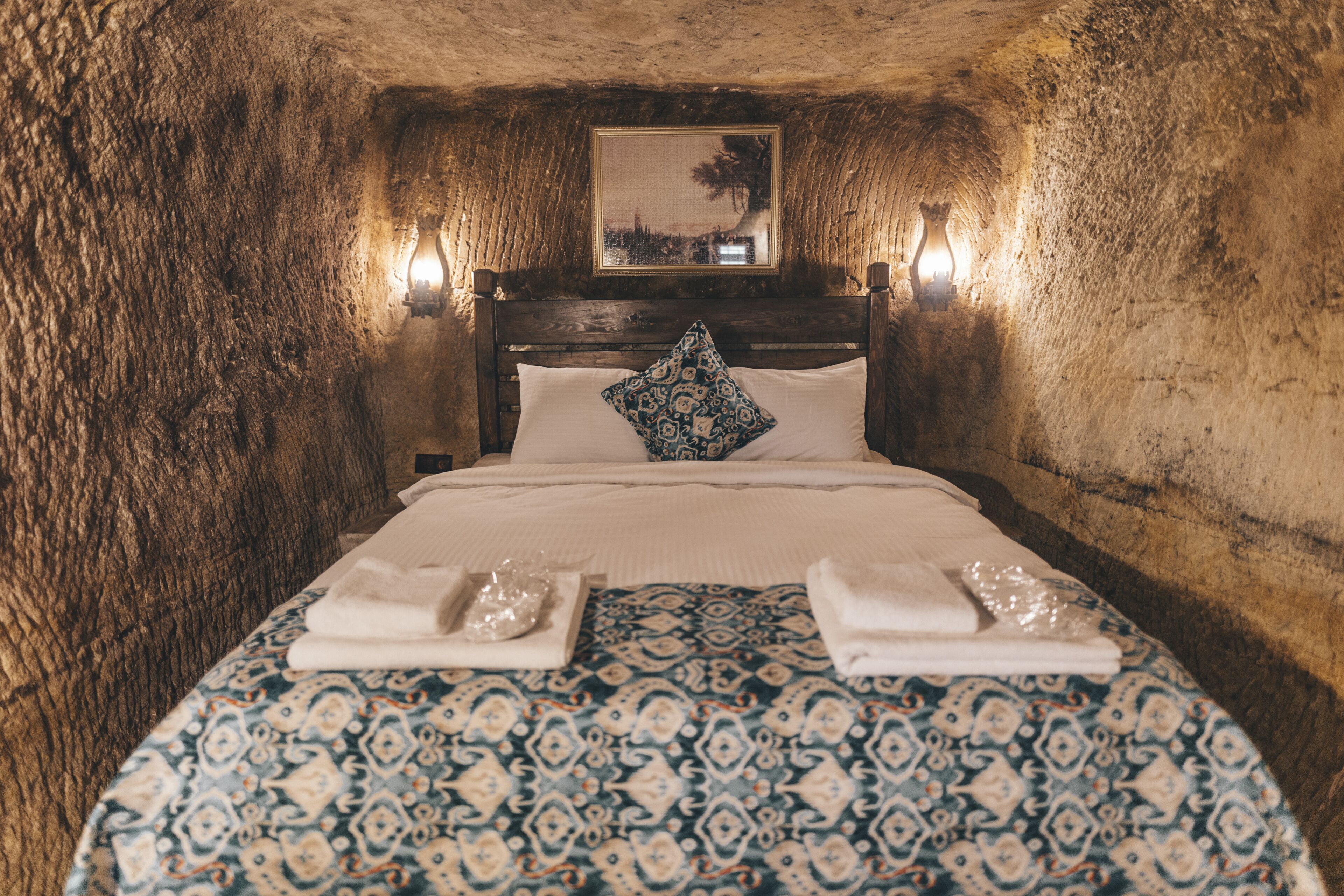 Una habitación con cama, cabecera de madera, edredón estampado y dos luces fijadas a la pared.