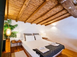 A bedroom with a wooden ceiling, a bed with white bedding, a bedside table with a lamp, and a potted plant.