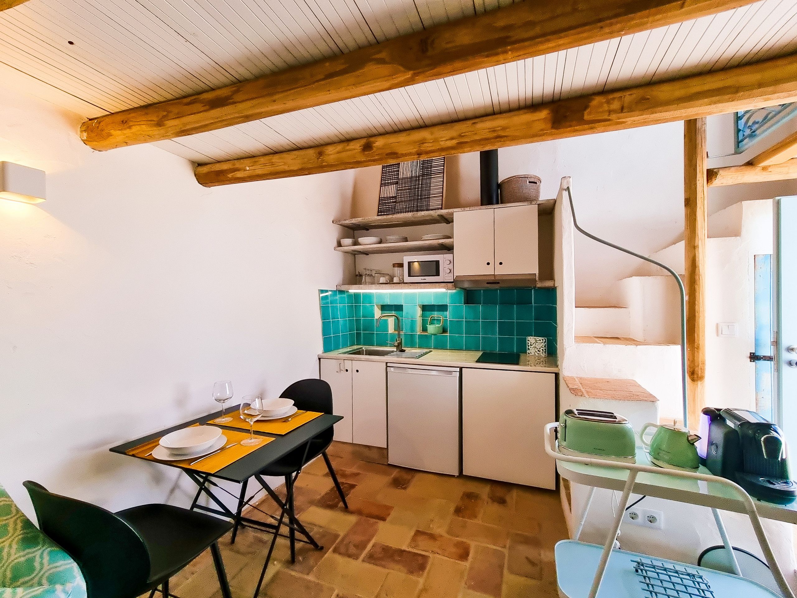 A compact kitchen with a microwave, sink, and cabinets. There is a dining table with chairs and a small table lamp.