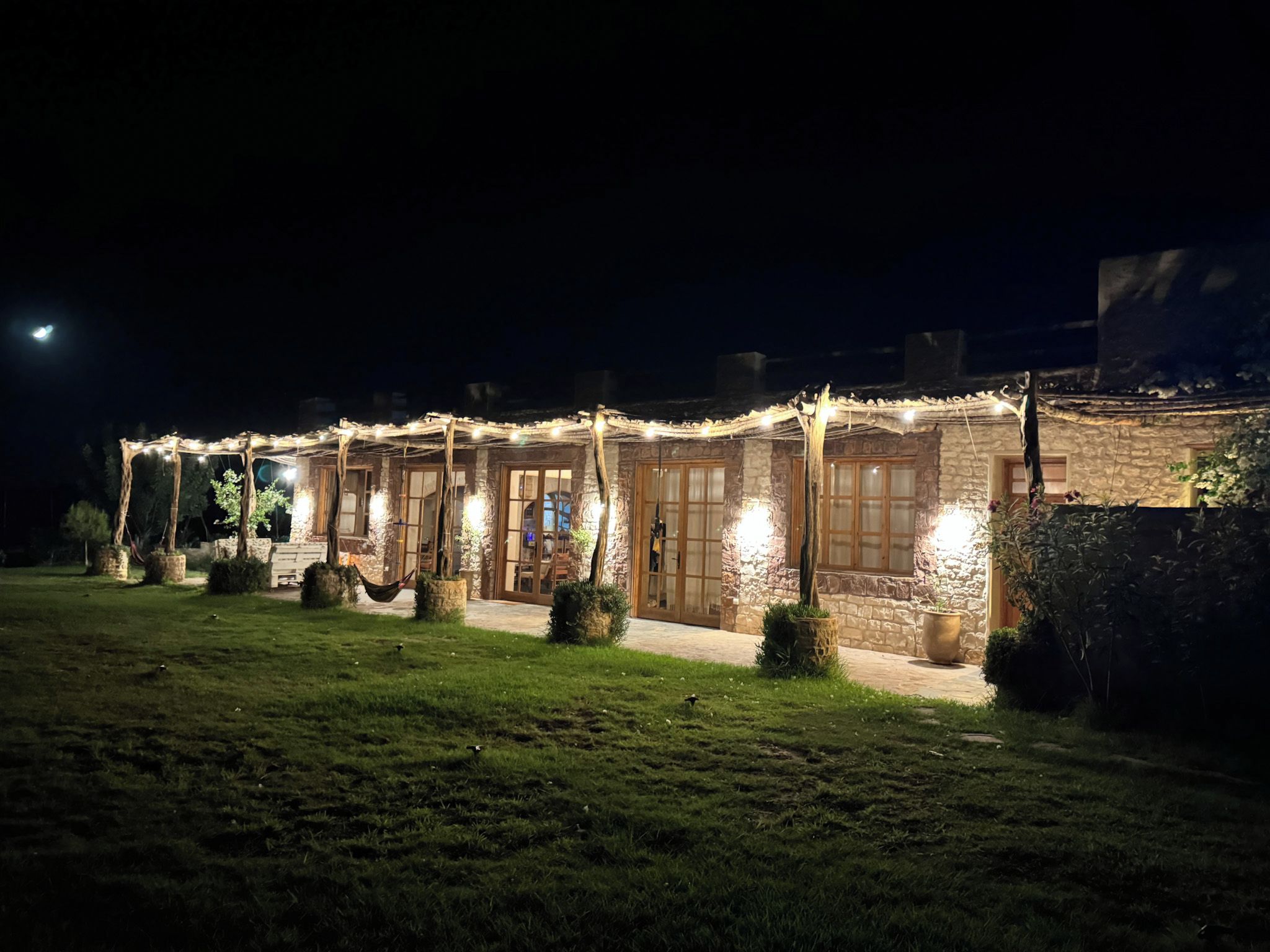 A stone building with multiple doors and windows, surrounded by a well-lit garden at night.