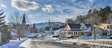 A snow-covered street with houses, a church steeple, and mountains in the background.