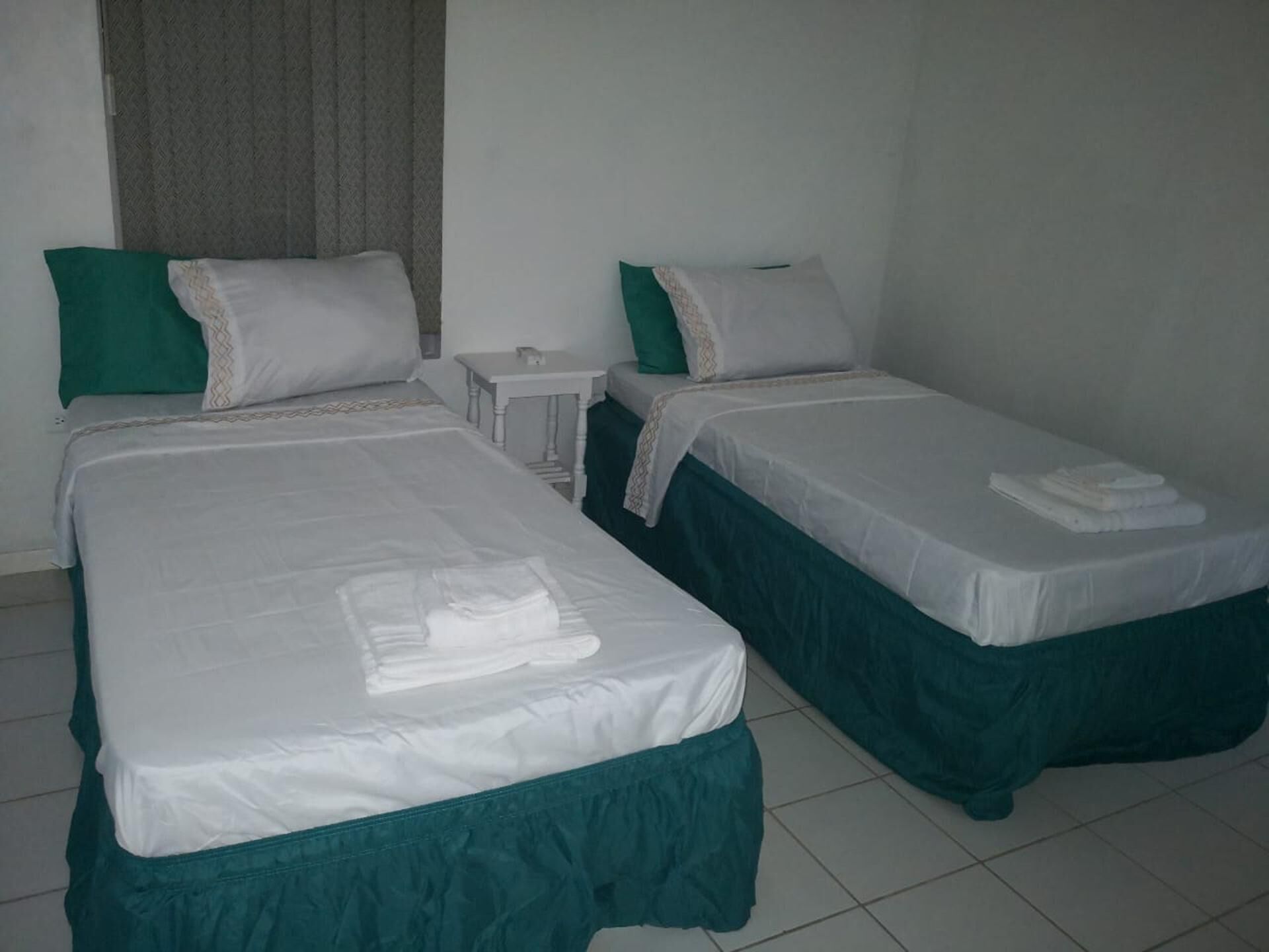 A hotel room with two single beds, each with a green bed skirt and white linens. There is a small bedside table with a lamp and a power outlet on the wall.