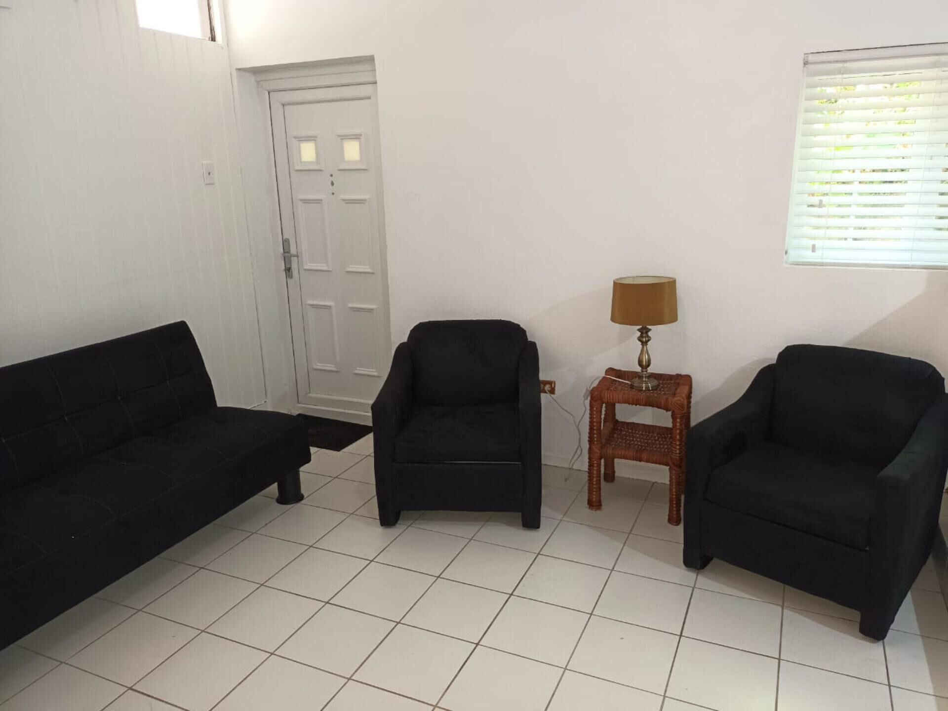 A room with two black sofas, a wooden side table with a lamp, a white door, and a window with blinds.