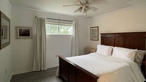 Un dormitorio con cama de madera, ventilador de techo, ventana con cortinas e imagen enmarcada en la pared.