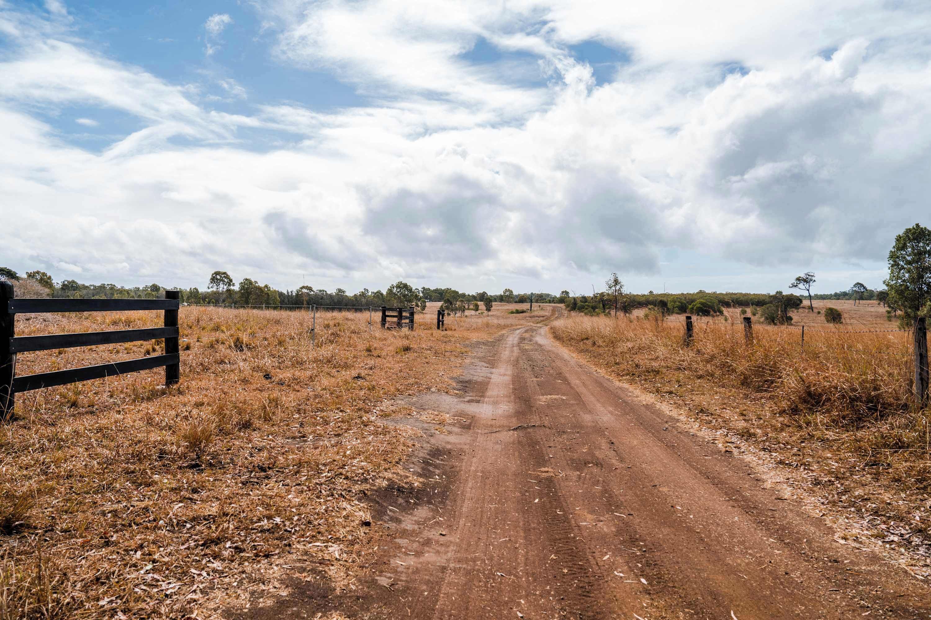 A dirt road leading through a dry, grassy field with a wooden fence on the left and trees in the distance.
