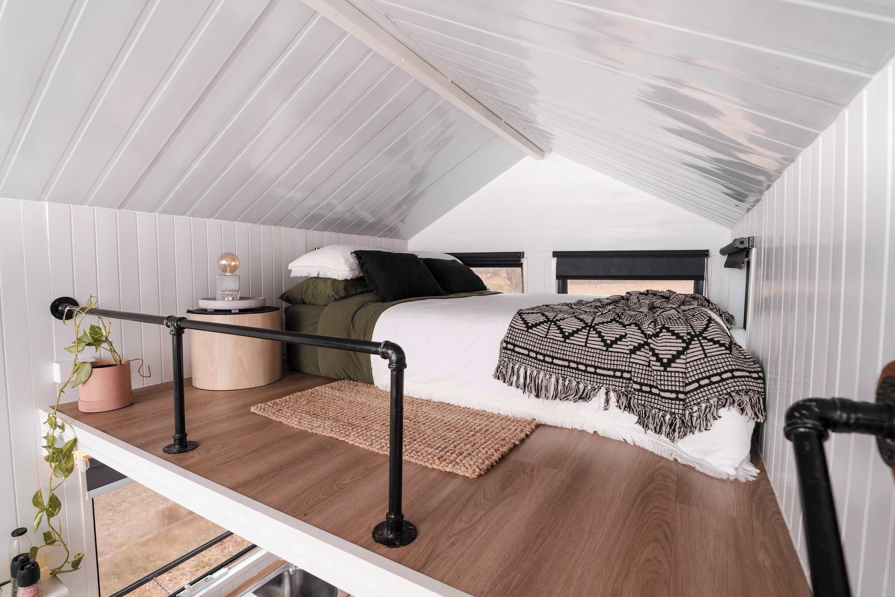 Dormitorio estilo loft con techo inclinado, cama con manta estampada, mesita de noche pequeña y una planta en maceta.