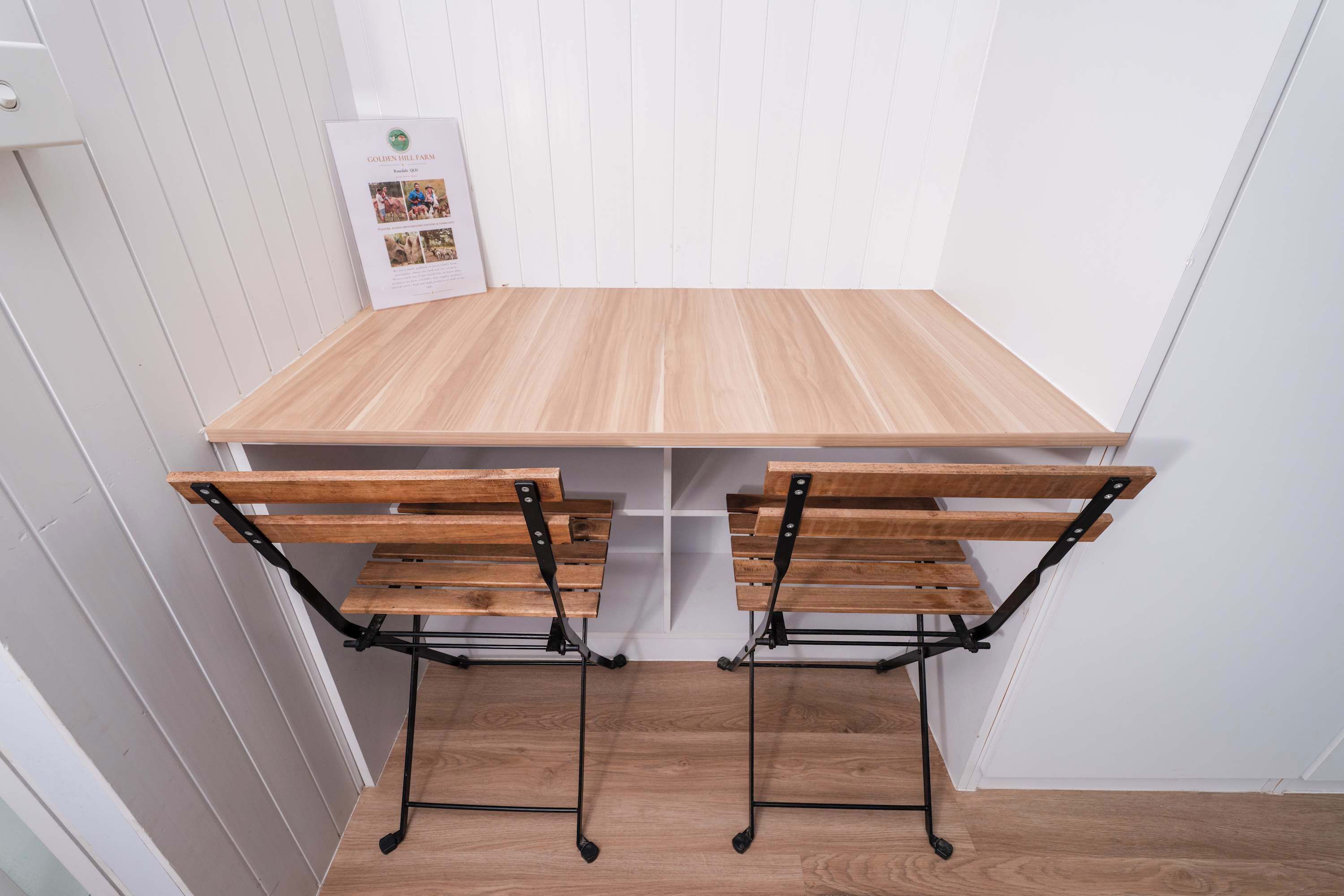 A wooden table with two chairs, a white wall with a bulletin board, and a wooden floor.
