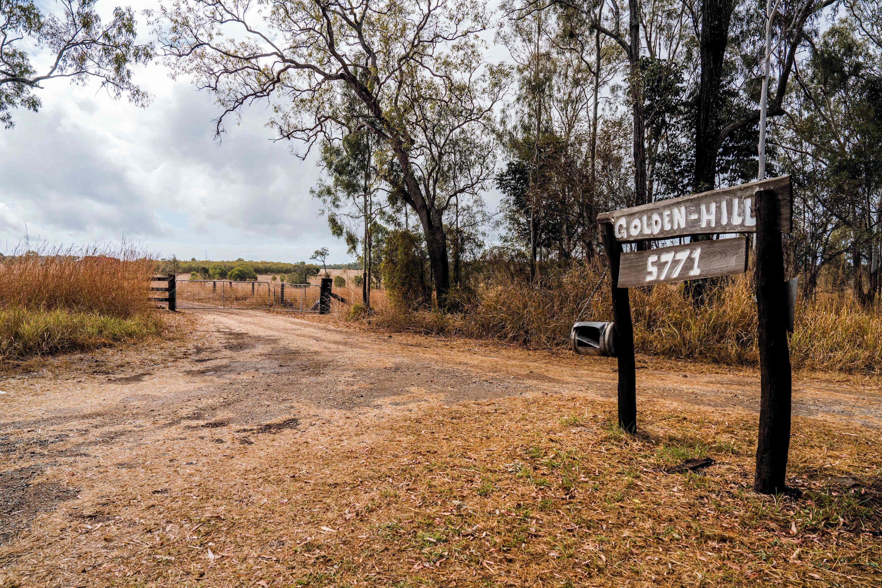 A dirt road leading to a wooden signpost with 'GOLDEN-HILL 5771' written on it, surrounded by trees and dry grass.