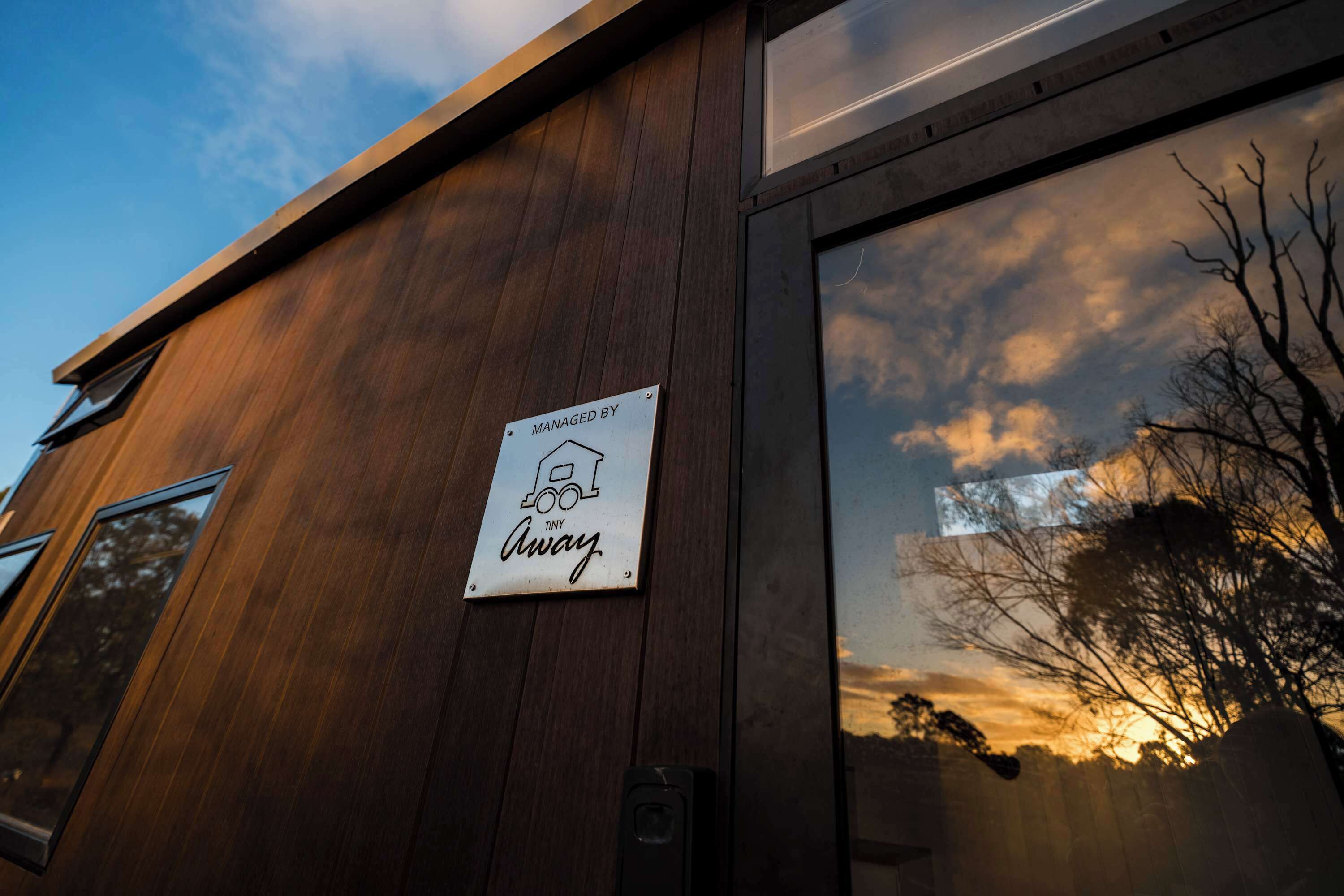 Ein Gebäude mit Holzfassade, ein Schild mit der Aufschrift 'Tiny Away' und ein Fenster, das einen Sonnenunterhimmel reflektiert.