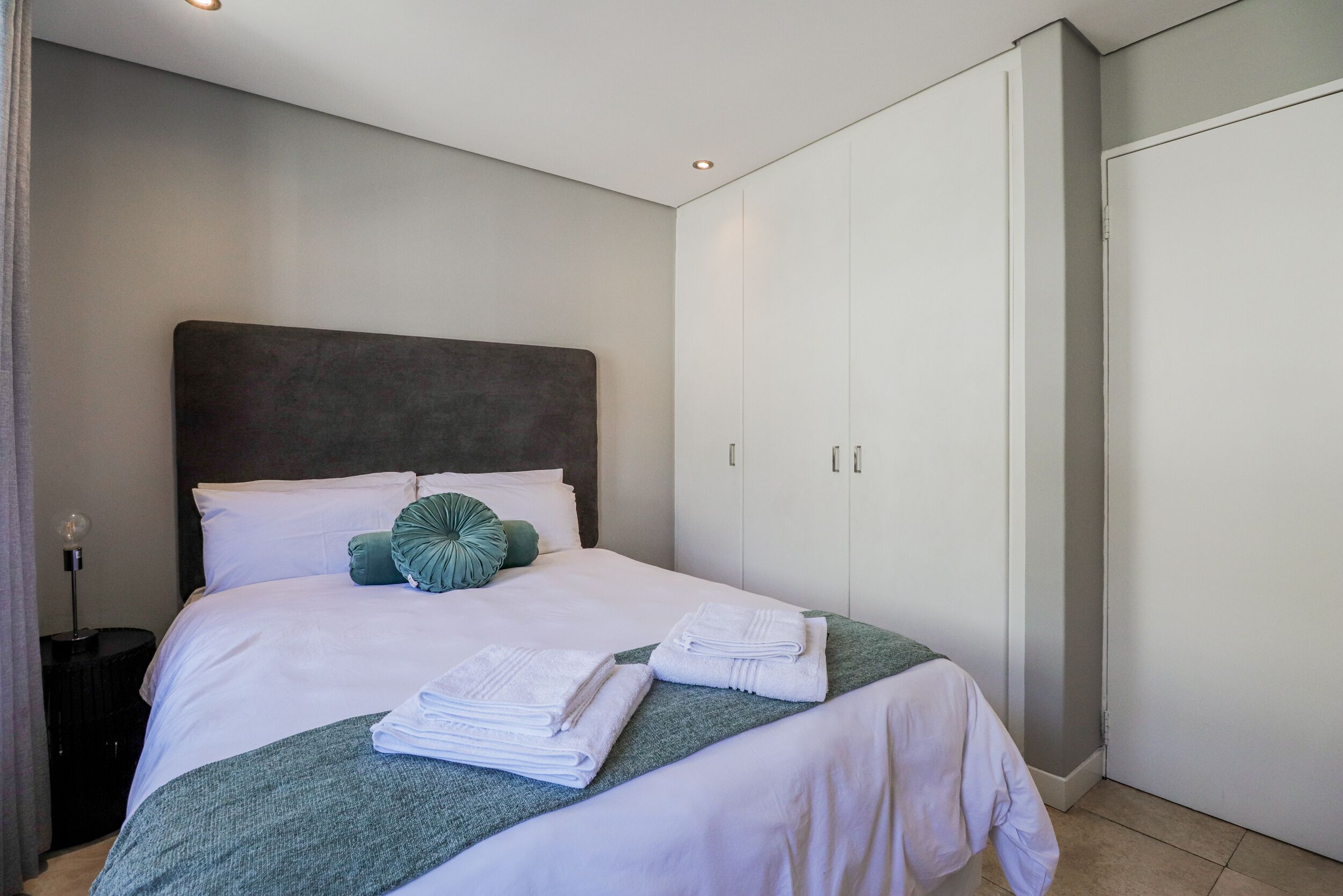 A neatly made bed with white linens and a green throw blanket, a bedside lamp, and a wardrobe with white doors.