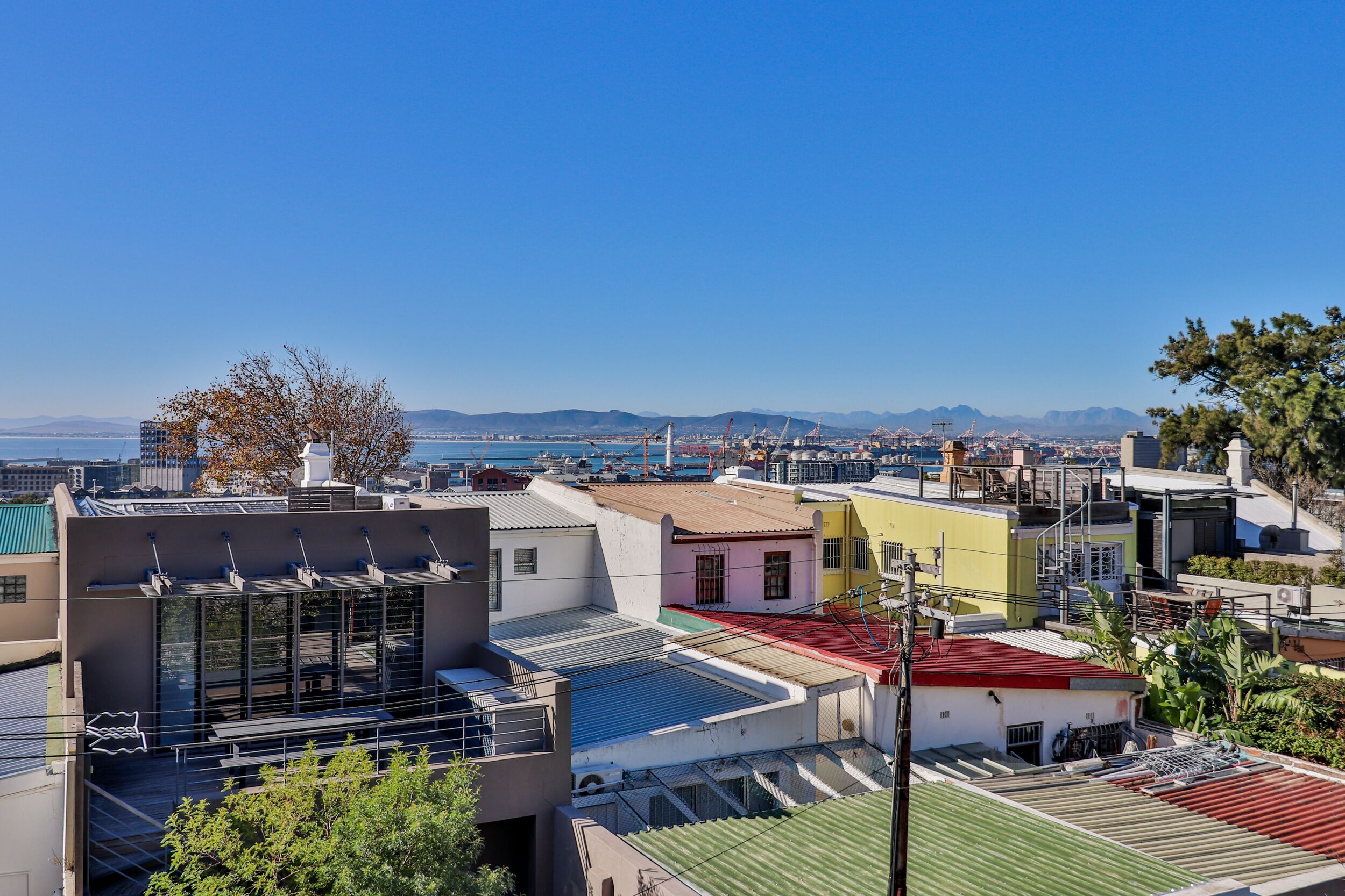 A coastal residential area with modern buildings, a clear blue sky, and a view of the sea and distant mountains.