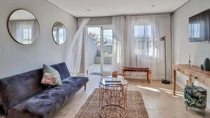 Una sala de estar moderna con un sofá, una mesa de centro y un televisor. Hay dos espejos en la pared, una ventana con cortinas y vistas al exterior.