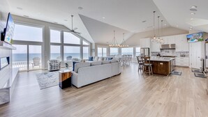A spacious living area with a large sofa, dining table, and chairs, a kitchen with modern appliances, and large windows offering ocean views.