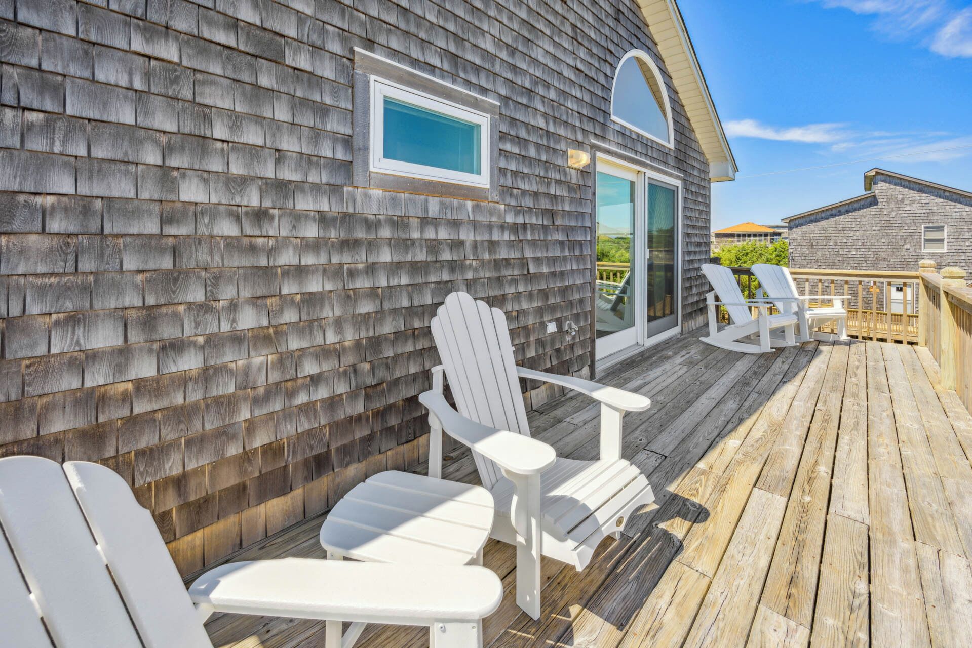Una terraza de madera con sillas Adirondack y un ventanal que da a un espacio cubierto.